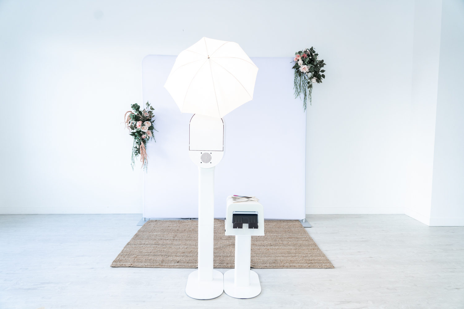 A DSLR photo booth is in front of a white backdrop. There is a flash on the top and to the right is a printer that prints the photo strips. The backdrop has floral arrangements on each corner of the white tension photo booth backdrop