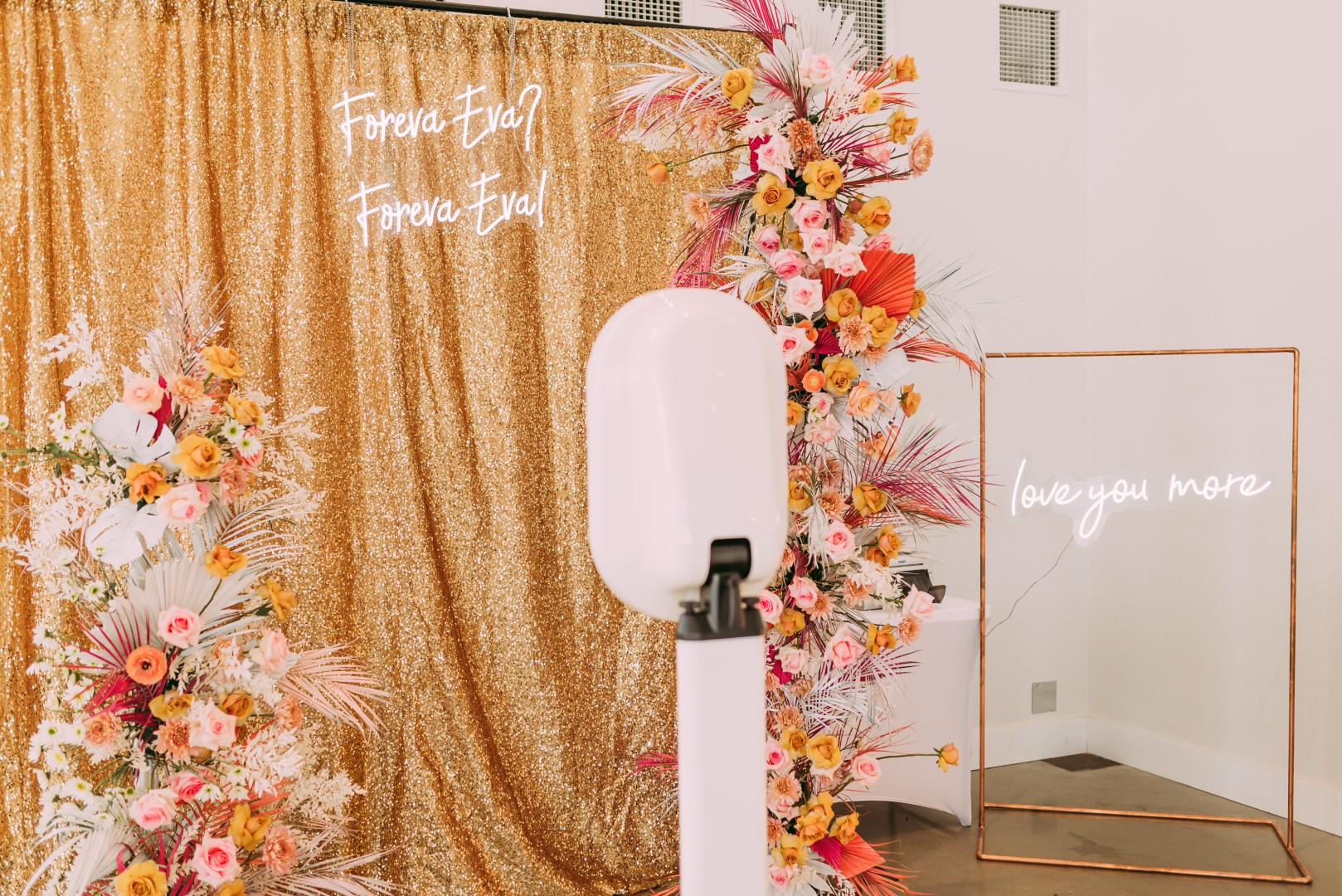 NWA Photo Booth is placed in front of a shimmery gold sequin backdrop and surrounded by bright prink and orange flowers at Osage House in Northwest Arkansas. To the right of NWA Photo Booth there is a neon sign in white light that reads I love you more, hanging from a copper pipe stand.