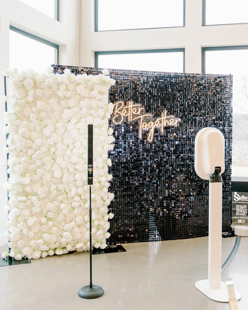 A white pedestal photo booth, provided by NWA Photo Booth, is in front of Stellar Party Co's black shimmer wall backdrop, layered with a white rose backdrop at Osage House

Large open windows are behind the backdrops letting in natural light to the Osage Reserve wedding venue.