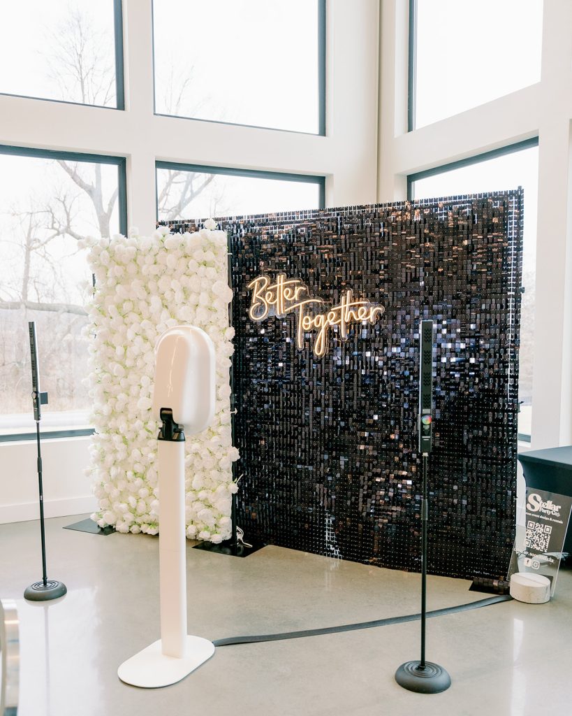 A white pedestal photo booth, provided by NWA Photo Booth, is in front of Stellar Party Co's black shimmer wall backdrop, layered with a white rose backdrop at Osage House

Large open windows are behind the backdrops letting in natural light to the Osage Reserve wedding venue.