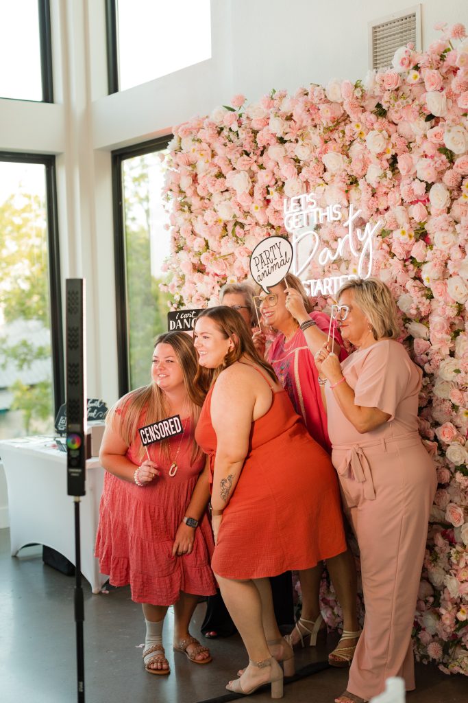 Wedding guests stand in front of a pink flower wall at Osage House while taking a photo with a pedestal photo booth from NWA Photo Booth. The wedding guests are dressed in fun bright reds and oranges to reflect the summer months of this wedding